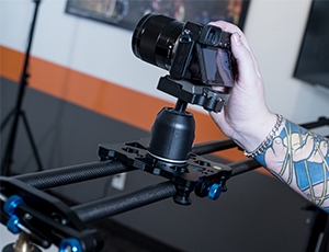 The tattooed arm of a content creator holds a mirrorless camera attached to a video ball head on a Cameron video slider.