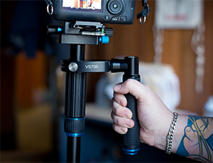 The tattooed arm of a content creator holds the handle on a Cameron video stabilizer with DSLR attached.