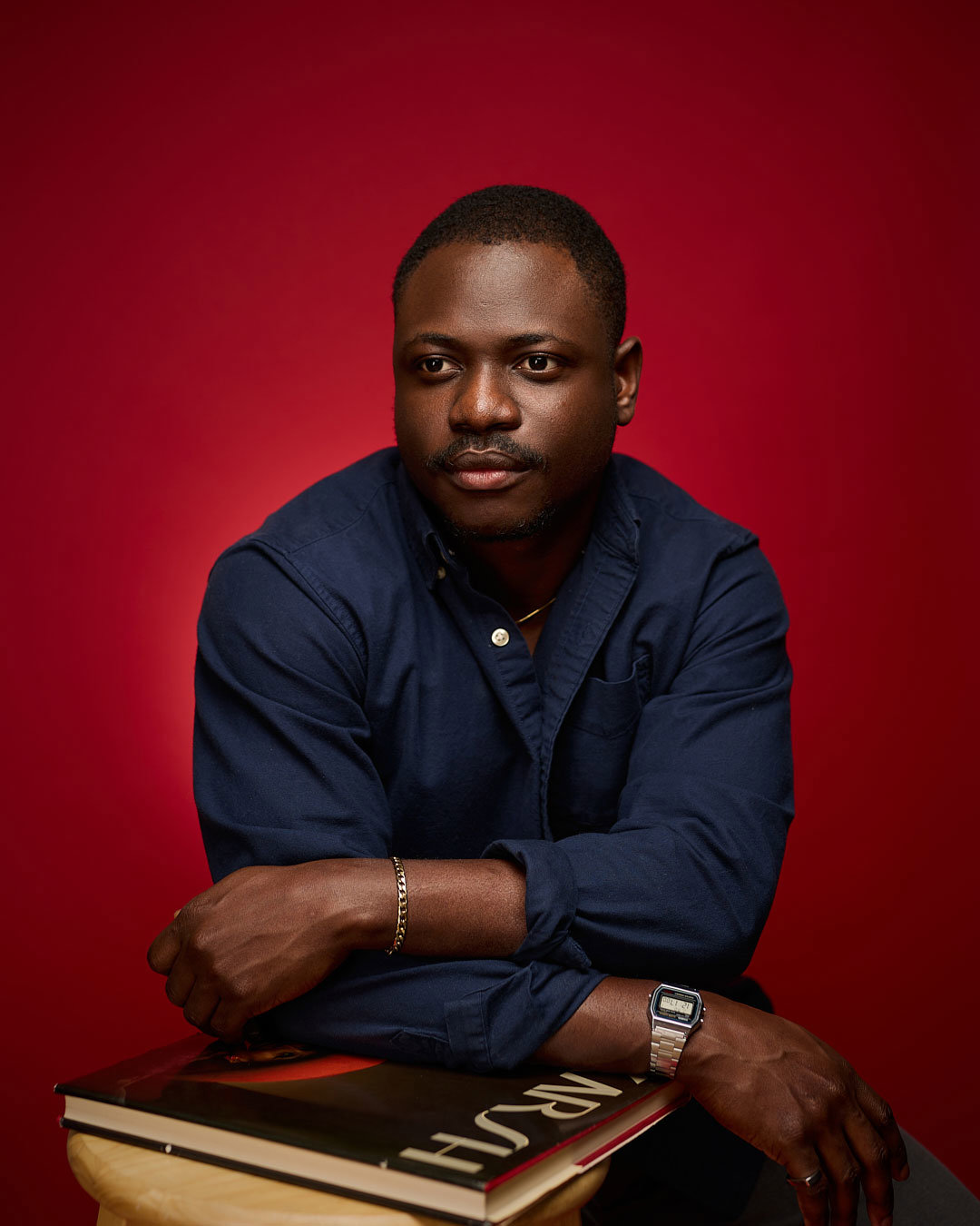 A young black man with short hair leaning forward with his arms over a book on a stool