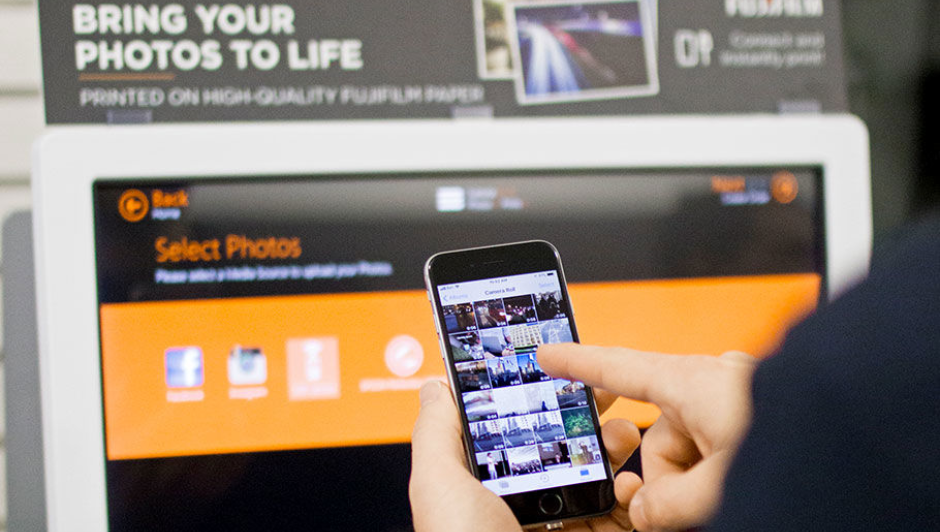 Close up of someone holding a mobile phone in front of a photo finishing kiosk