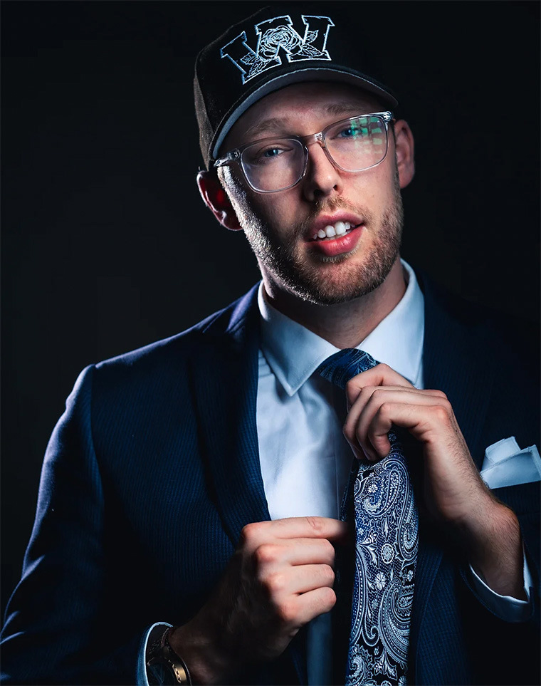 A young man wearing glasses, a suit, and a baseball cap
