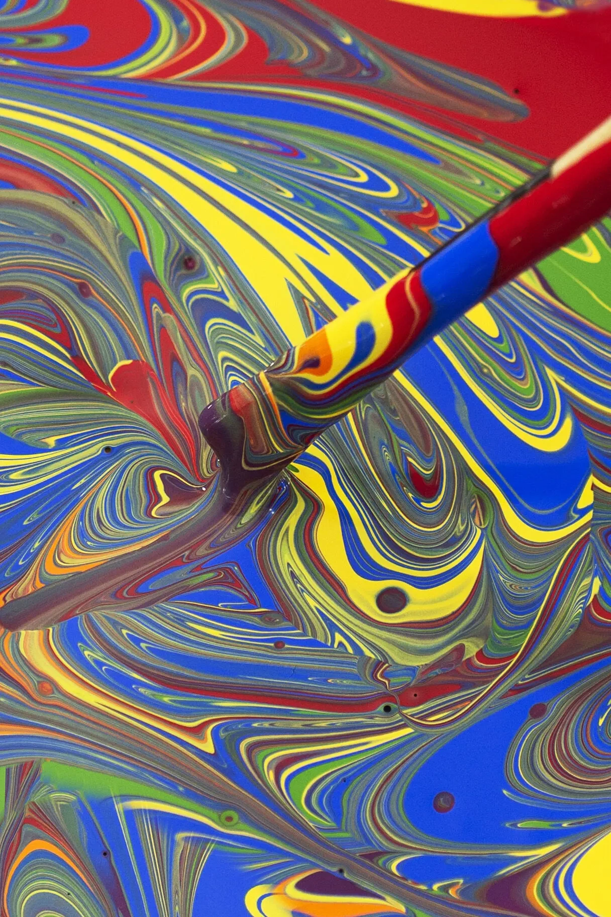 Macro photo of someone blending swirls of red, yellow, blue and green paint with a stick