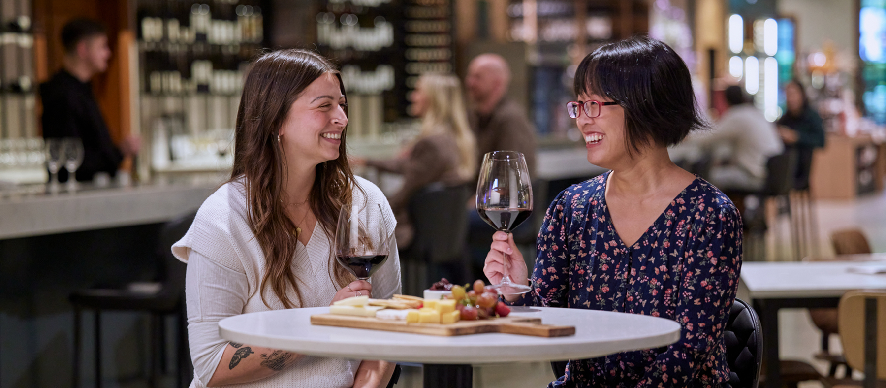 Two women enjoying red wine while eating charcuterie with the tasting bar in the background