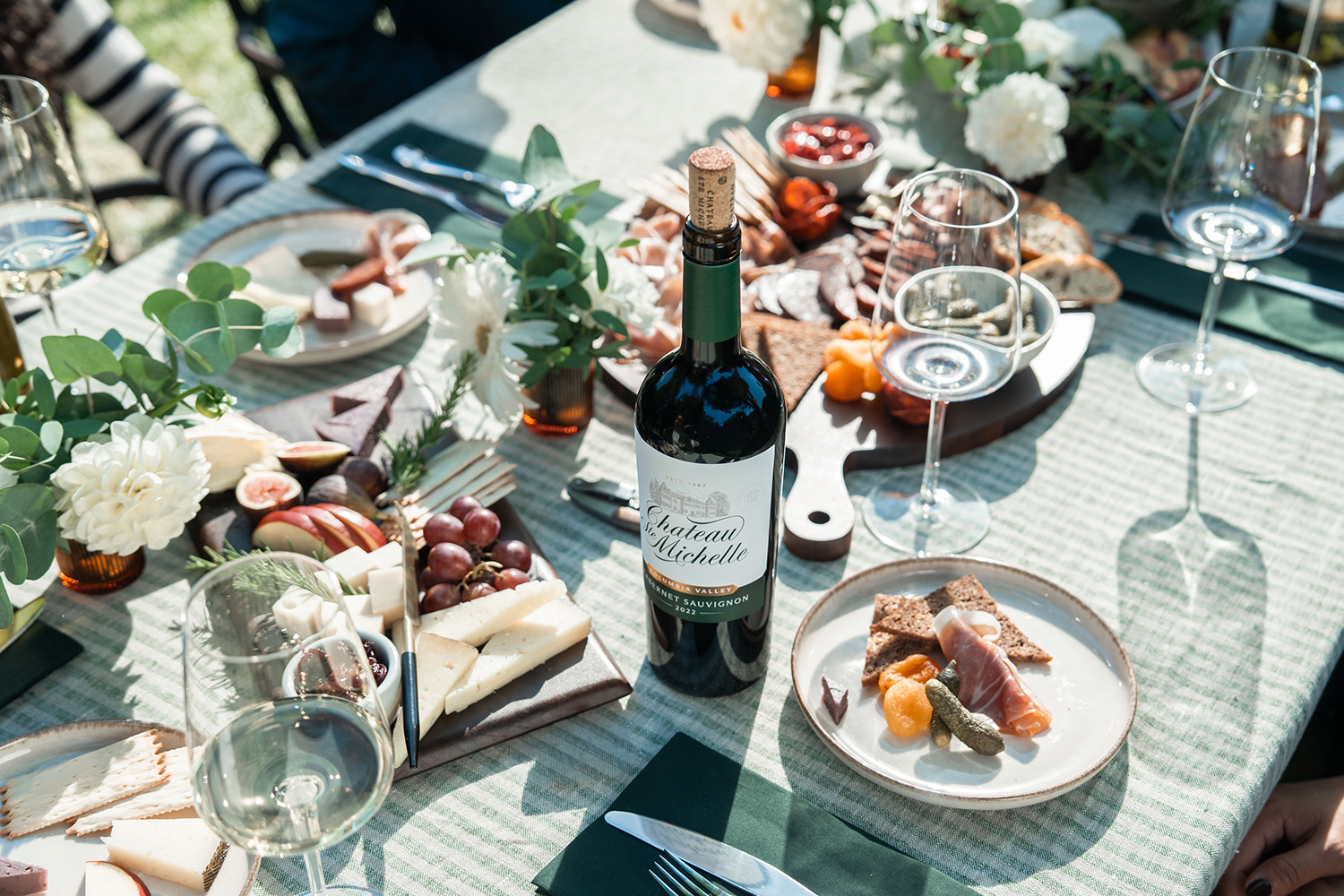 A bottle of Chateau Ste Michelle Cabernet Sauvignon on a table with a charcuterie board and white wine