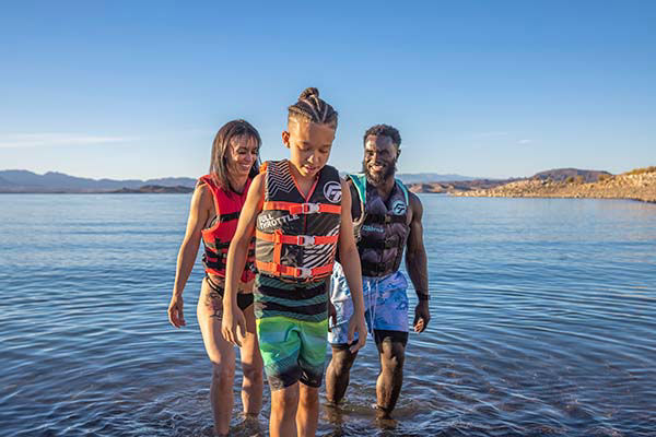 A family in life jackets walks through shallow water.