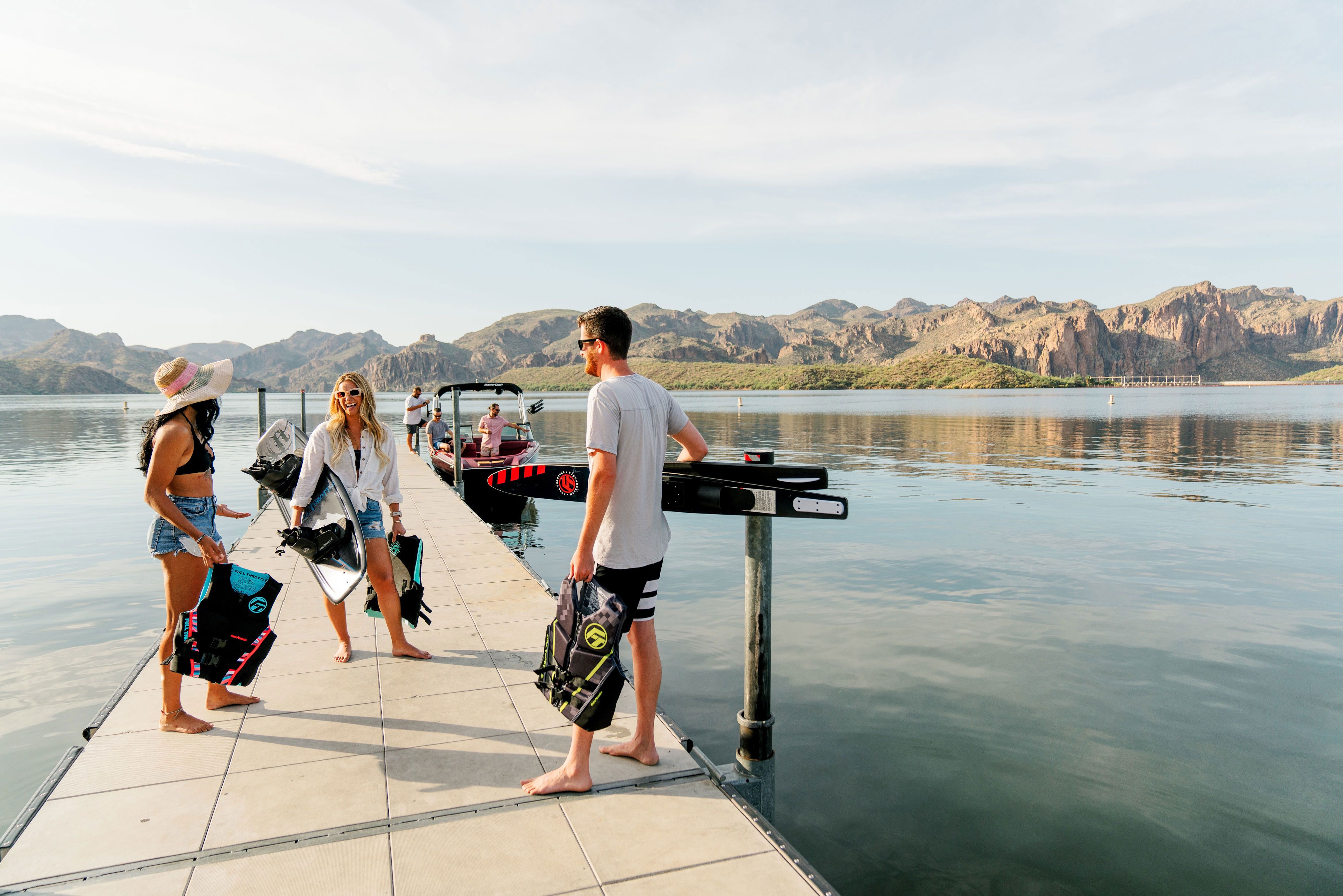 Three friends stand on a dock near the water, holding boating gear.
