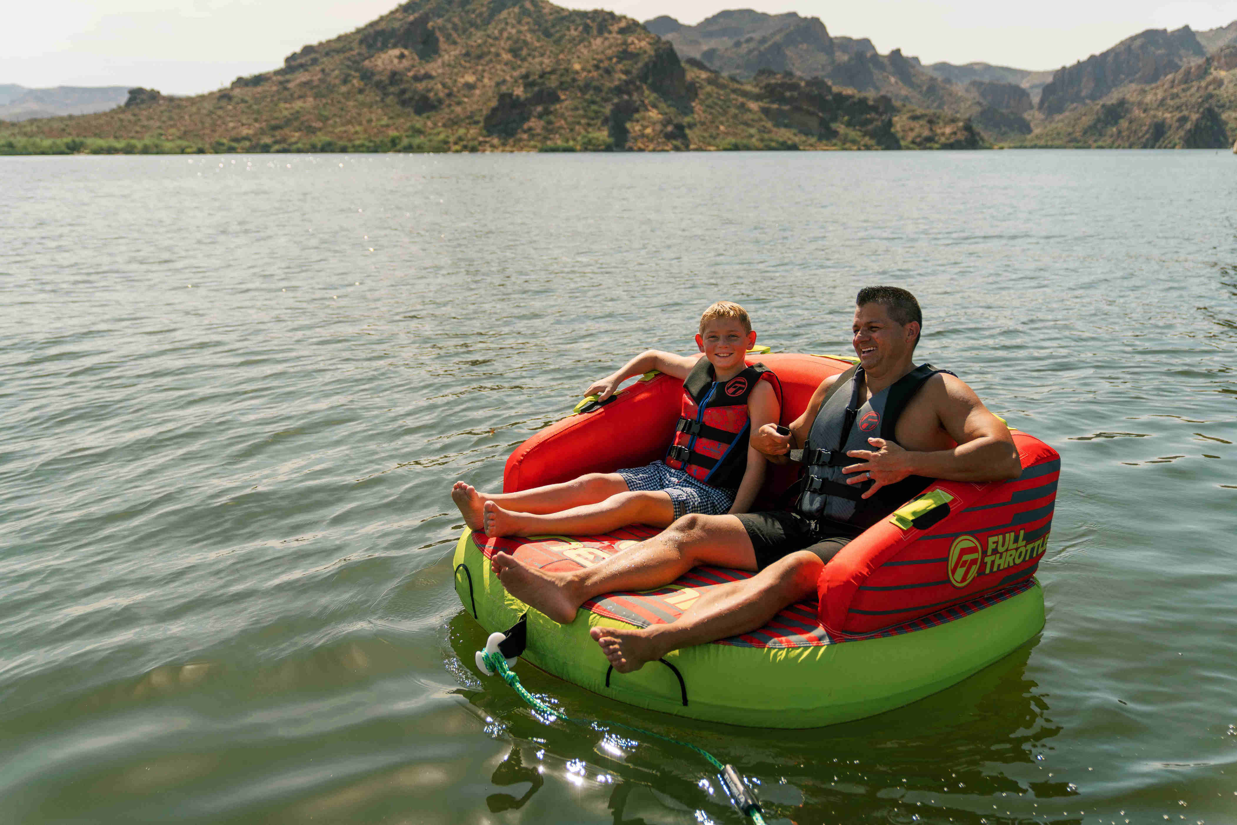A father and son sit in a towable tube on the water with hills in the background.