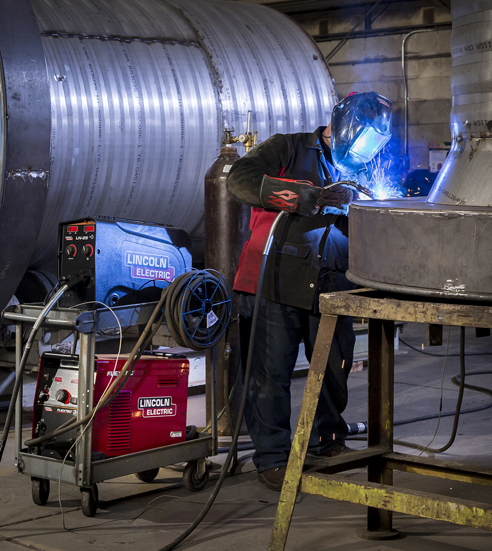 Worker performing a weld on a piece of industrial machinery using Lincoln Electric welding equipment