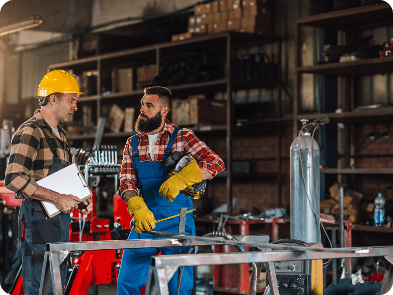 a man in welding gear speaking with a man in a hard hat in a workshop