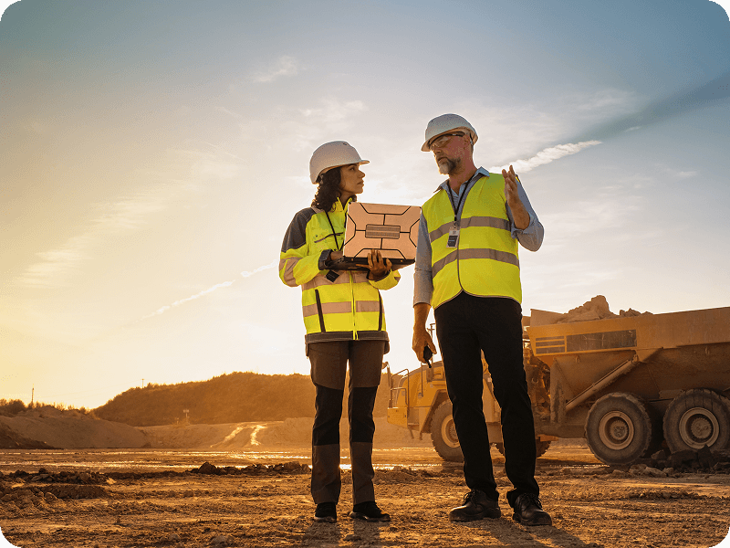 a man and a woman on a sandy construciton site going over something on a laptop